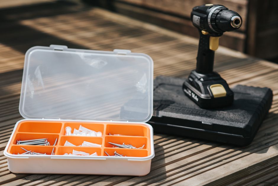 A black cordless drill-driver standing on its case beside an open box of screws and wall plugs on decking