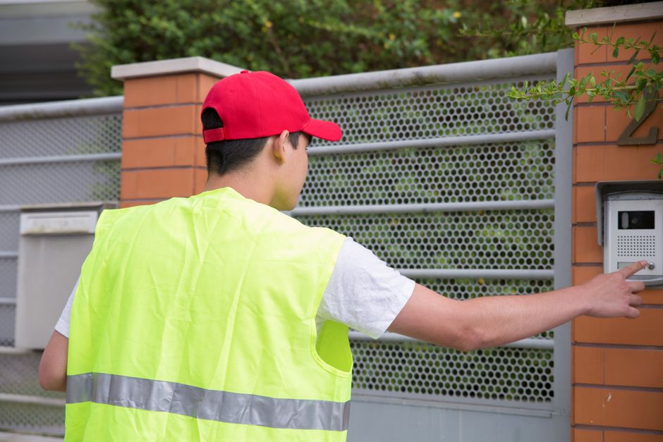 A courier in a hi-vis vest pressing a camera doorbell on a brick gatepost