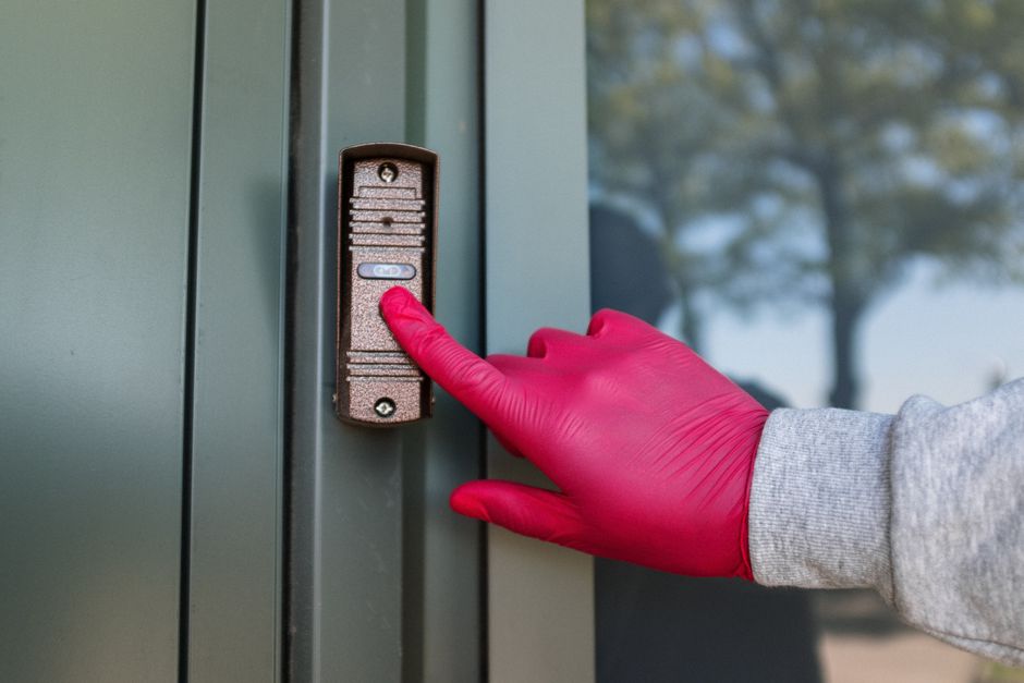 A gloved hand pressing a wall-mounted doorbell unit with a small camera lens on a grey metal door frame