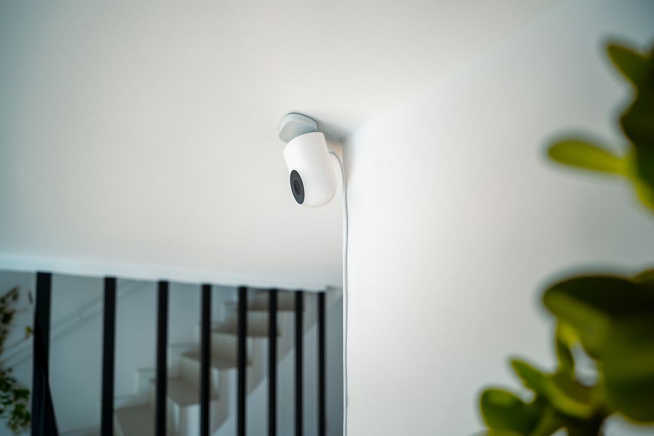 A small white wired security camera mounted high on a wall above a staircase