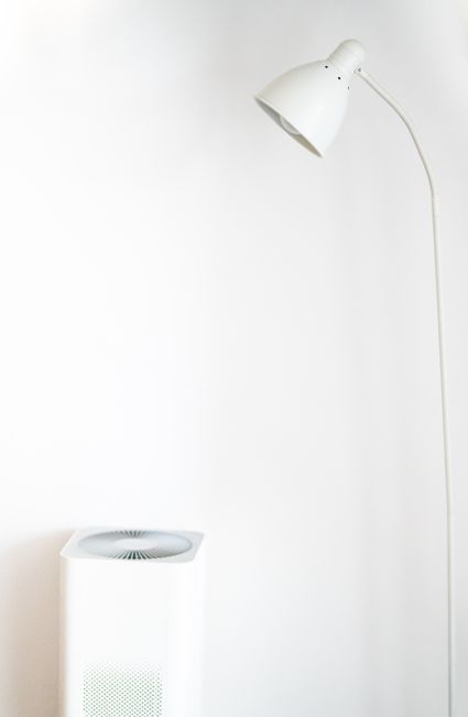 A white air purifier with a round top grille beside a white floor lamp against a white wall