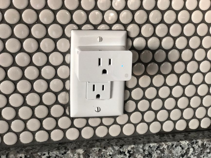A white smart plug with a blue status light plugged into a wall outlet on a penny-tile splashback