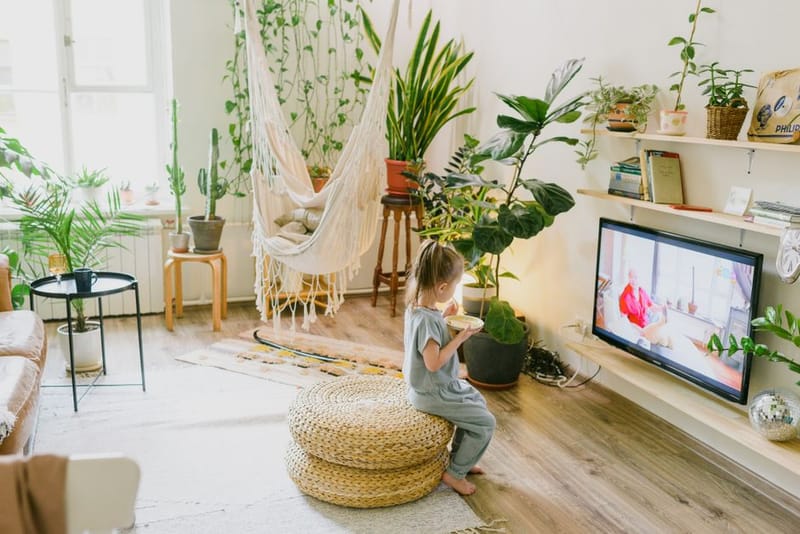 A small child on a woven pouffe watching TV in a bright living room full of plants